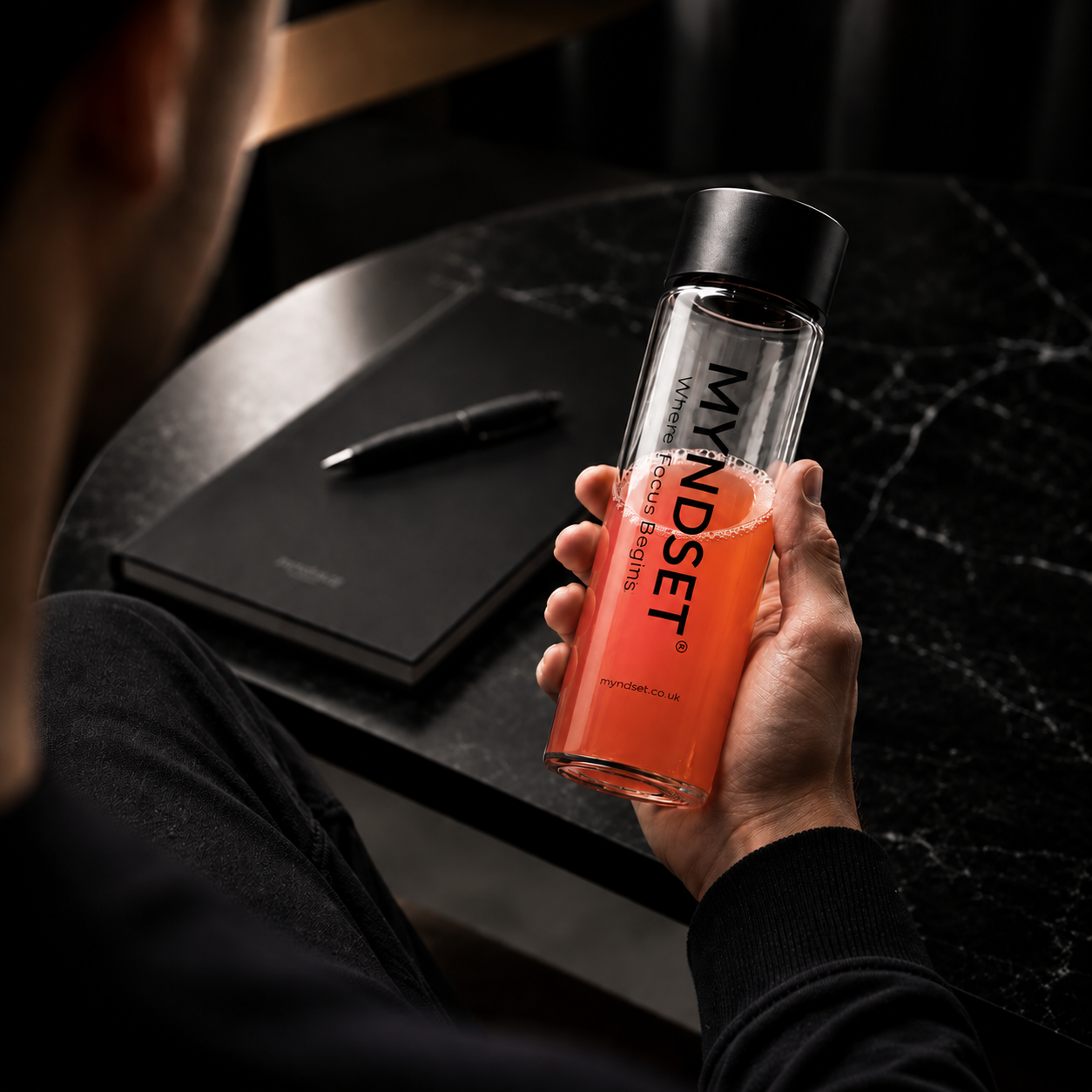 Person holding a glass bottle with a red liquid and 'MYNDSET' branding, sitting at a desk.