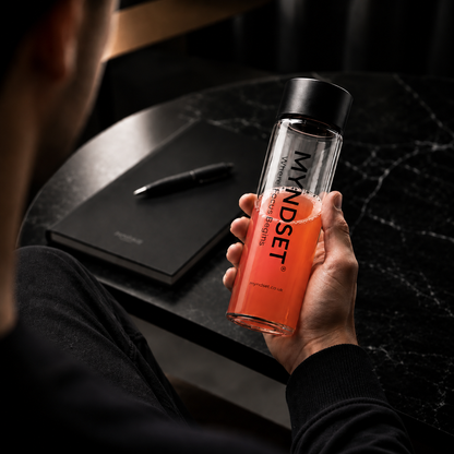 Person holding a glass bottle with a red liquid and 'MYNDSET' branding, sitting at a desk.